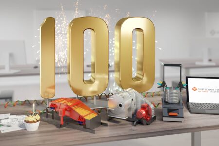 100 Jahre SIEBTECHNIK TEMA, Büro mit Schreibtisch auf dem eine große goldene 100 steht, im davor sind Modelle einiger Maschinen, eine konstruktionsskizze und ein Laptop. Bei den Maschinen handelt es sich um eine Zentrifuge, eine Siebmaschine und eine Analysensiebmaschine