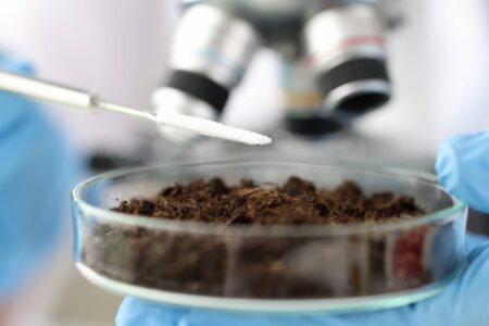 A laboratory employee holds a soil sample in a glass container in front of a microscope and adds white powder