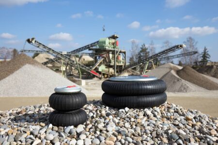 Two individual, differently sized air bellows of the LuCoTec air suspension system lie on a pile of gravel in a gravel works, in the background you can see the plant with screening machines and conveyor system